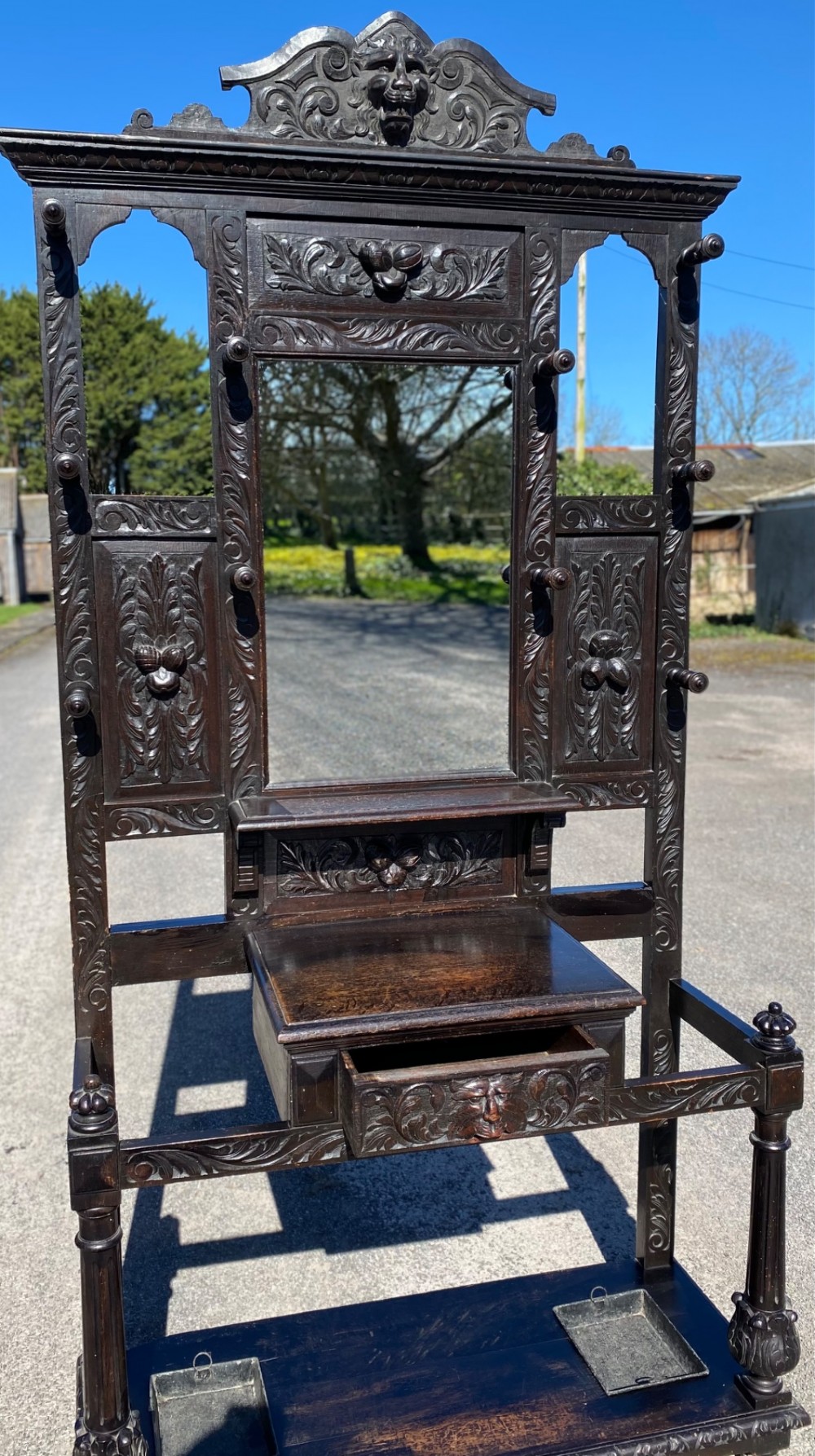 A dark carved oak Victorian hall stand featuring a central rectangular mirror, lion mask carvings, turned coat pegs, umbrella holders, and metal drip trays. View VII