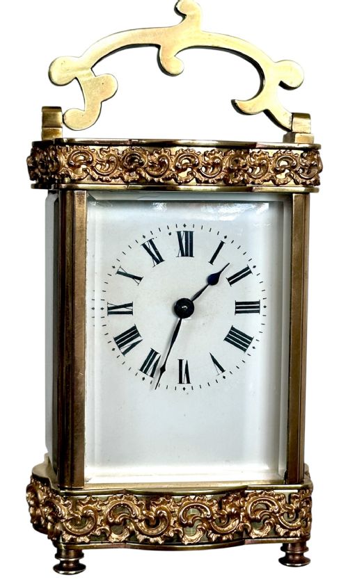 fig 1. An antique French brass carriage clock featuring serpentine glass panels, scrollwork borders, a white enamel dial with Roman numerals, and a carrying handle.