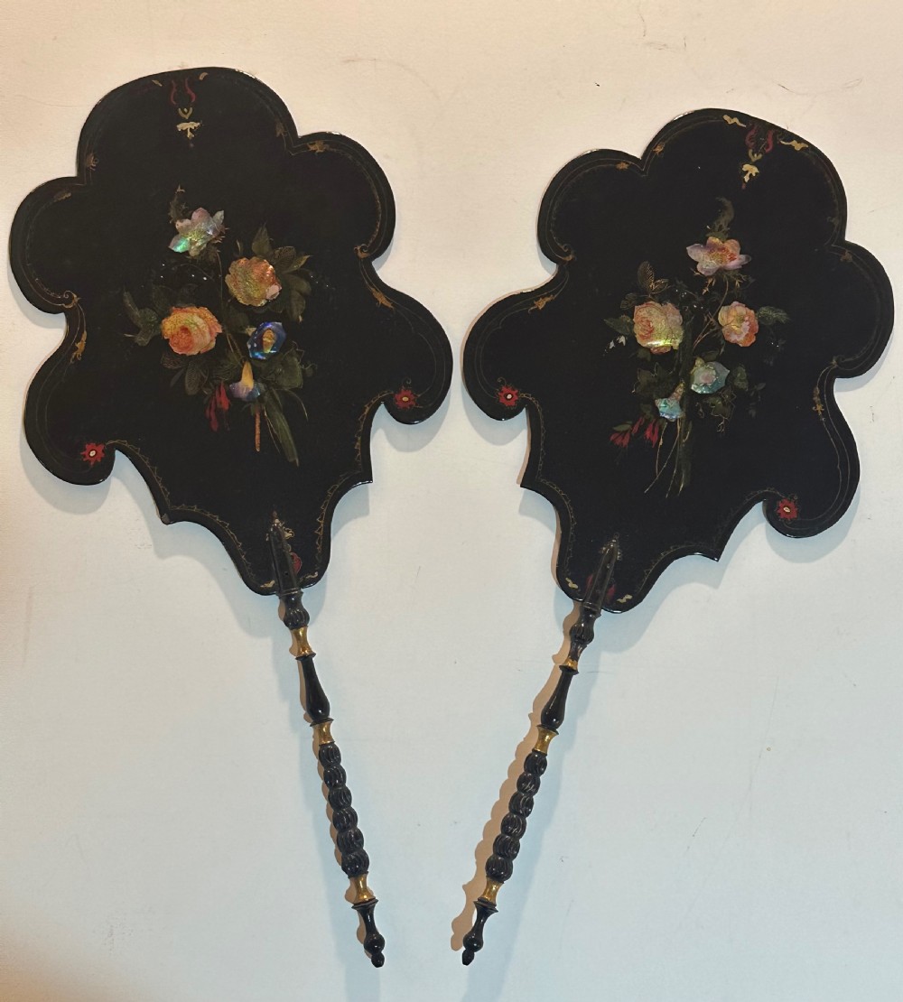 A pair of black Victorian papier-mache face screens with scalloped edges, hand-painted floral motifs inlaid with mother-of-pearl, and turned ebonised handles.