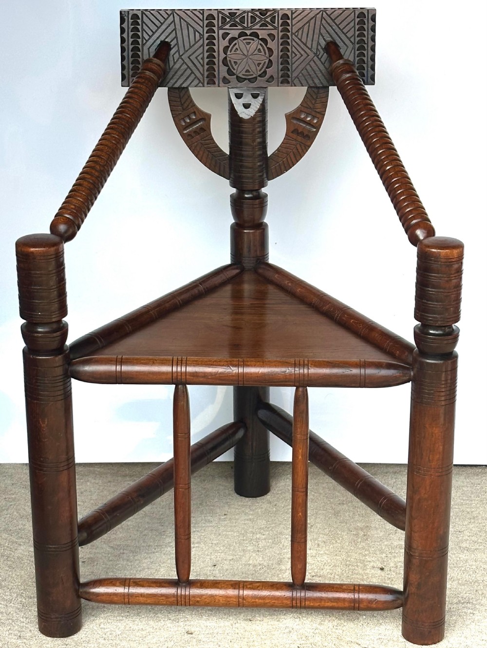 A dark oak three-legged turned chair featuring a triangular wooden seat, turned spindle arms, bobbin legs, and a carved geometric backrest panel.