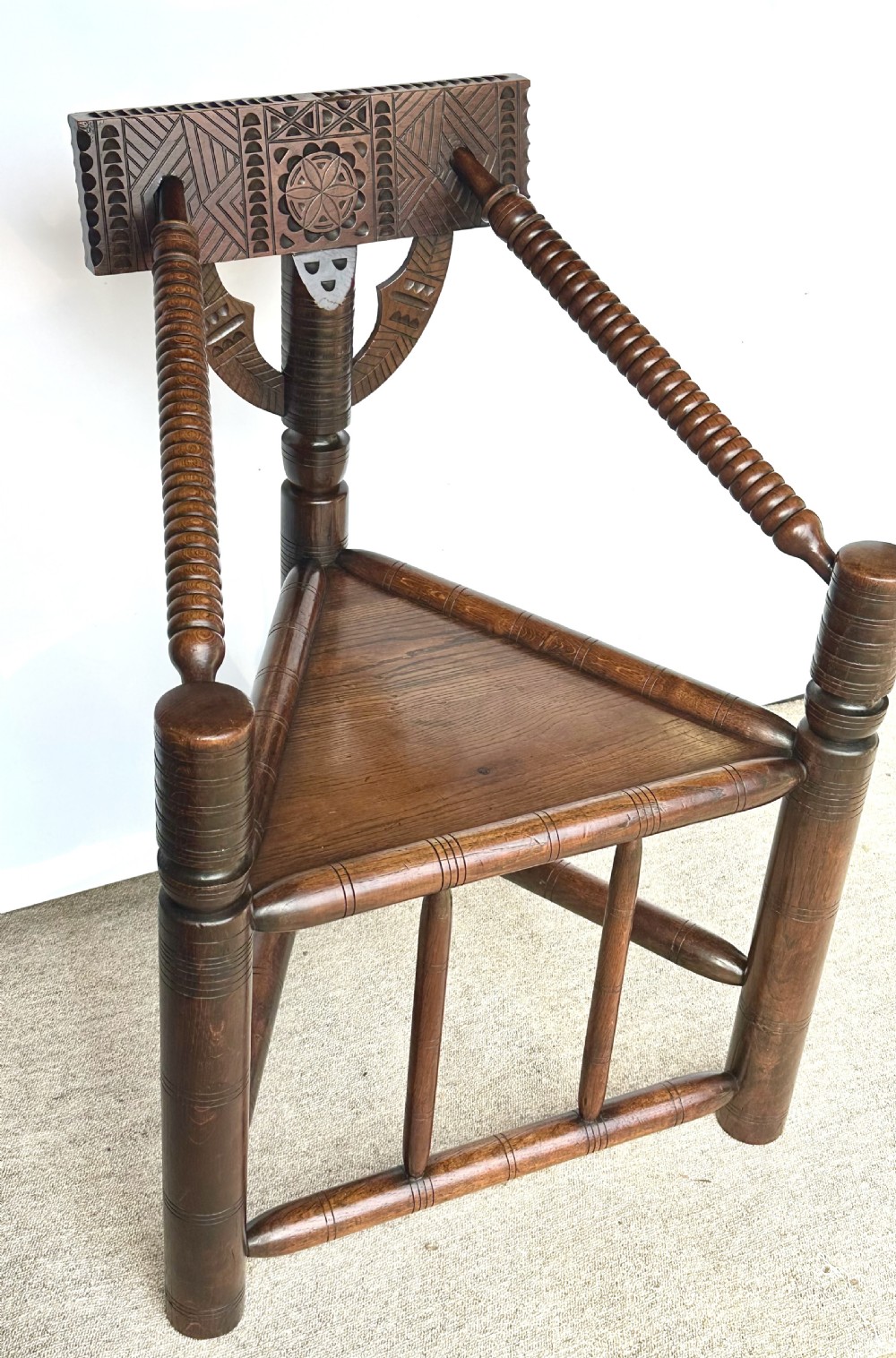 A dark oak three-legged turned chair featuring a triangular wooden seat, turned spindle arms, bobbin legs, and a carved geometric backrest panel. View II