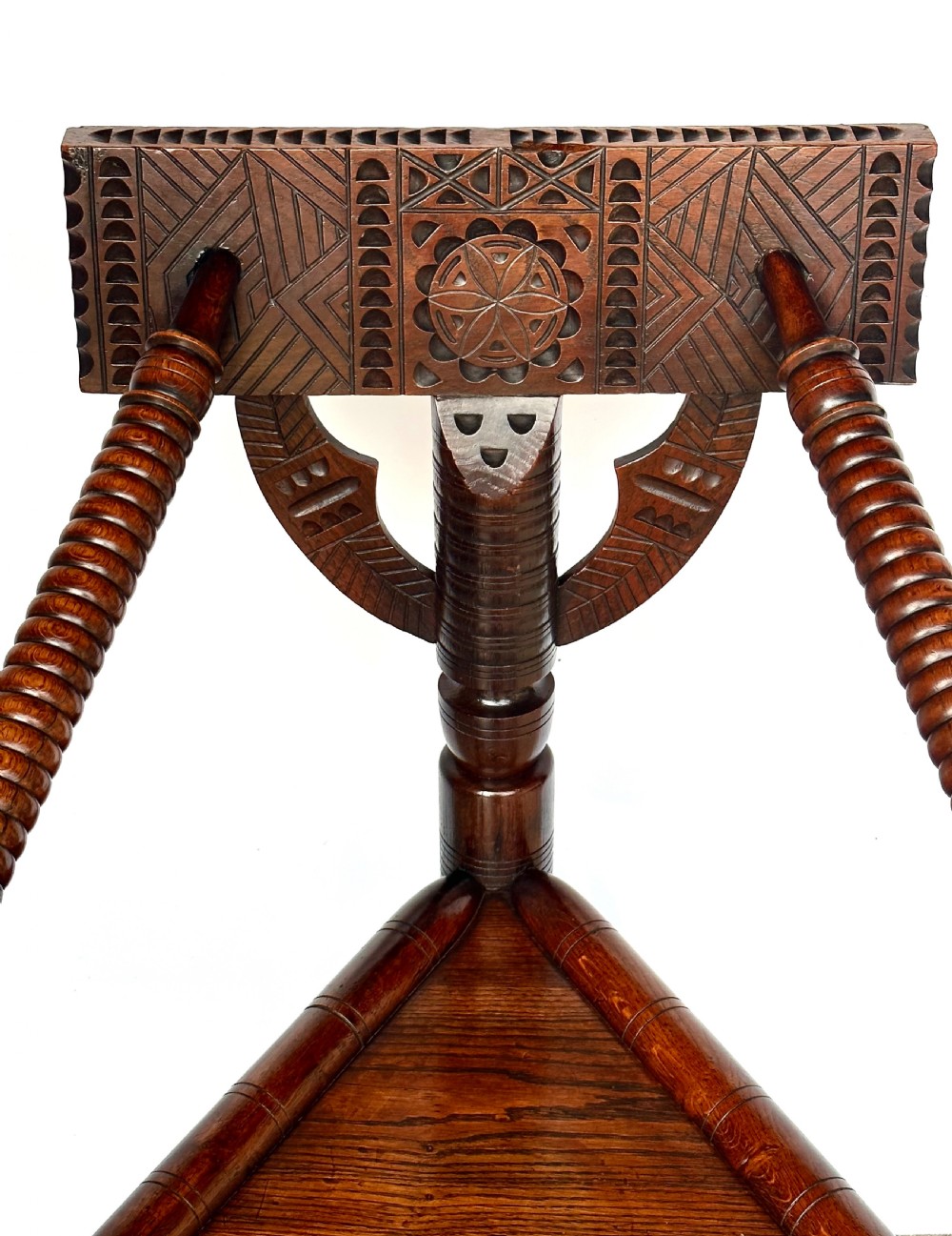 A dark oak three-legged turned chair featuring a triangular wooden seat, turned spindle arms, bobbin legs, and a carved geometric backrest panel. View IV
