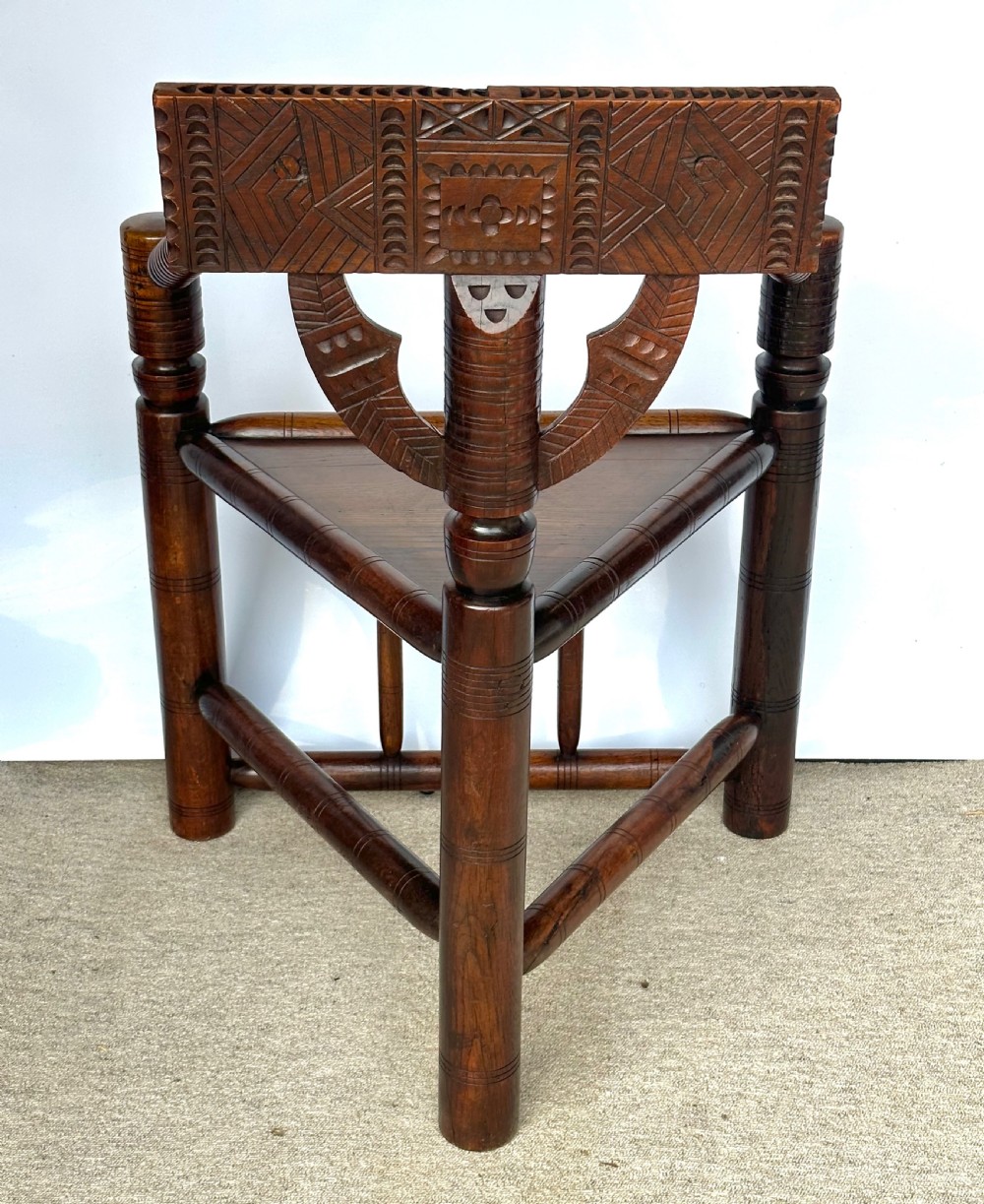 A dark oak three-legged turned chair featuring a triangular wooden seat, turned spindle arms, bobbin legs, and a carved geometric backrest panel. View V