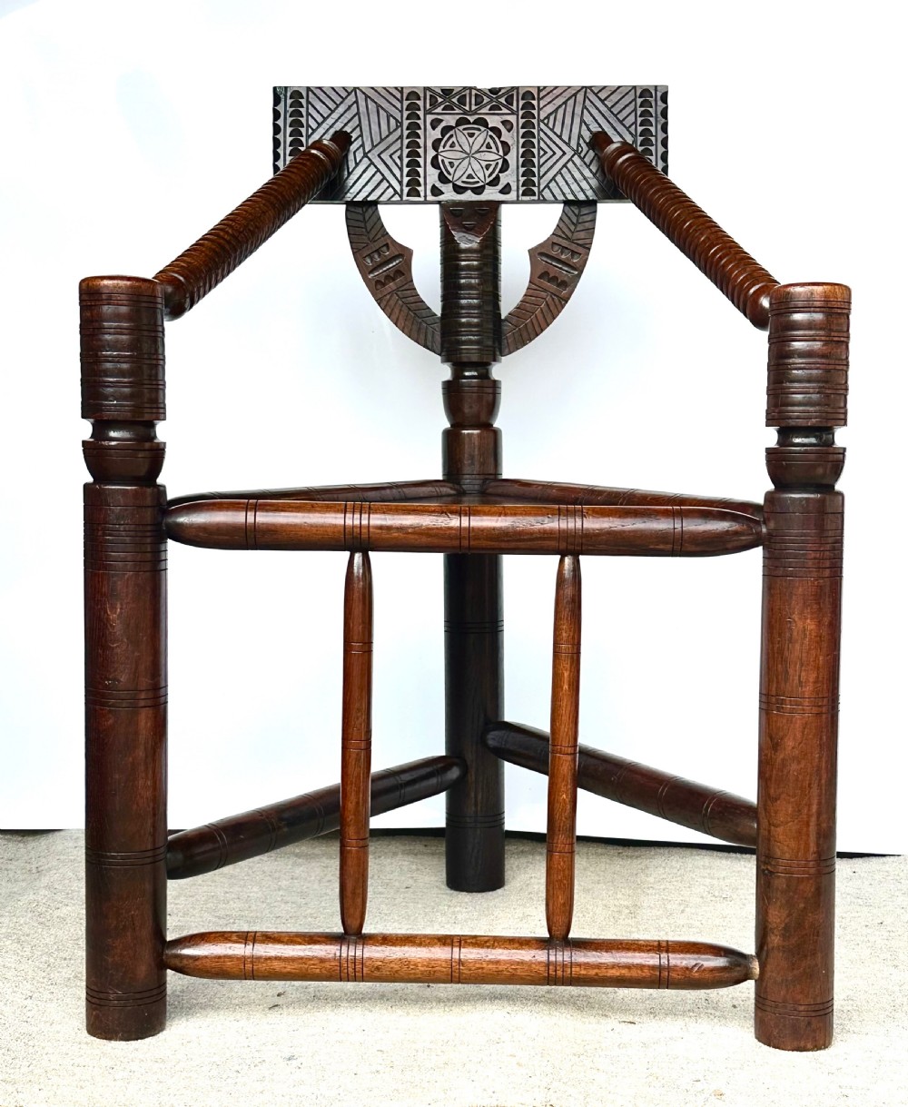 A dark oak three-legged turned chair featuring a triangular wooden seat, turned spindle arms, bobbin legs, and a carved geometric backrest panel. View VI