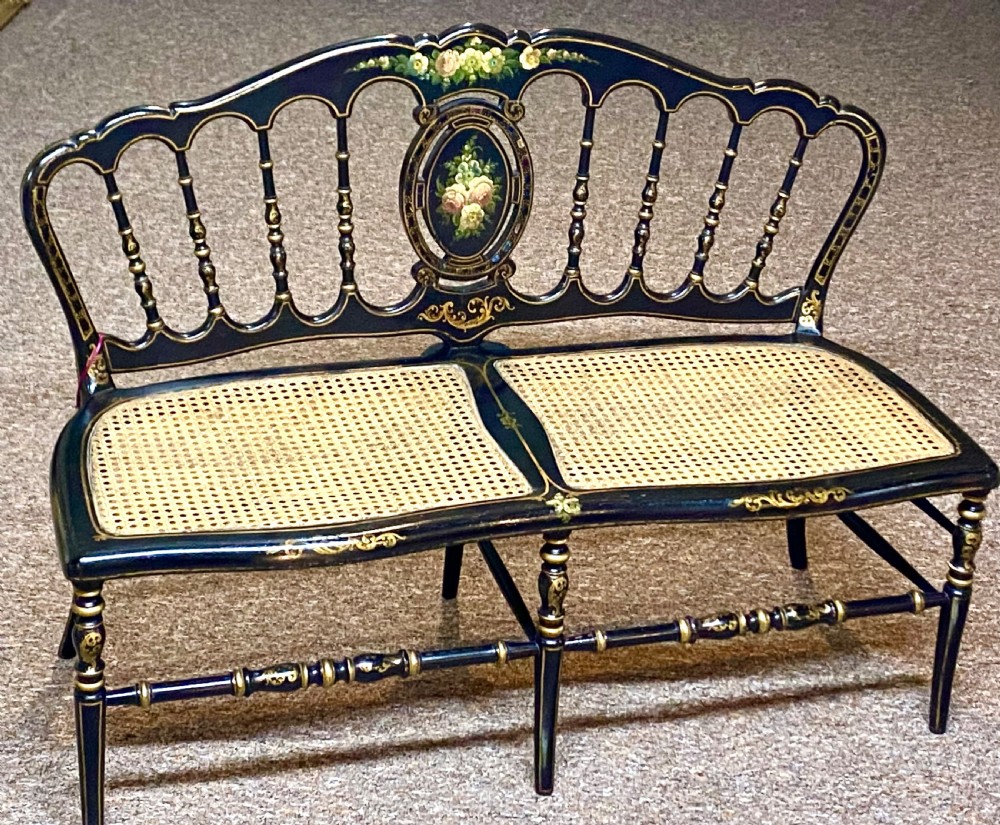 An antique black ebonised wooden bench with hand-painted floral panels, gold accents, turned spindle backrest, and two cane seat panels.