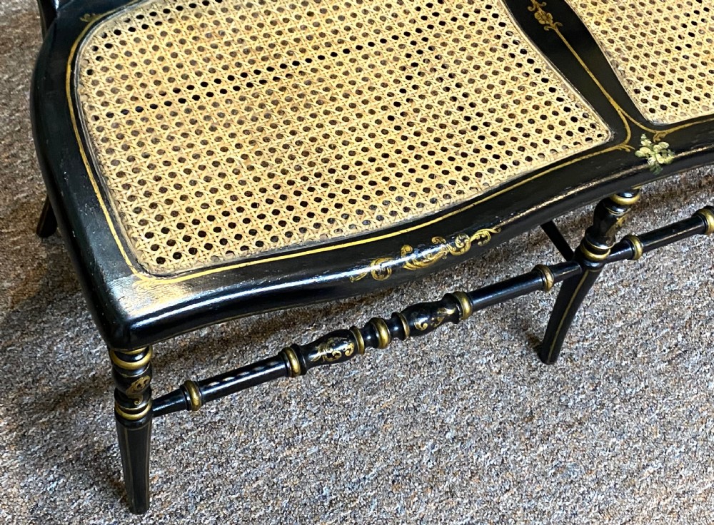 An antique black ebonised wooden bench with hand-painted floral panels, gold accents, turned spindle backrest, and two cane seat panels. View VII