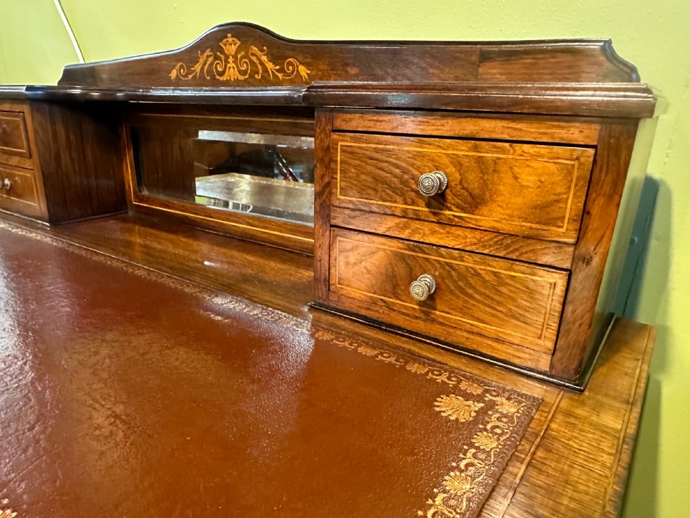 An Edwardian rosewood writing desk with satinwood marquetry, a dark brown leather writing surface, a raised superstructure with small drawers and a mirror, standing on tapered legs with castors. View VI