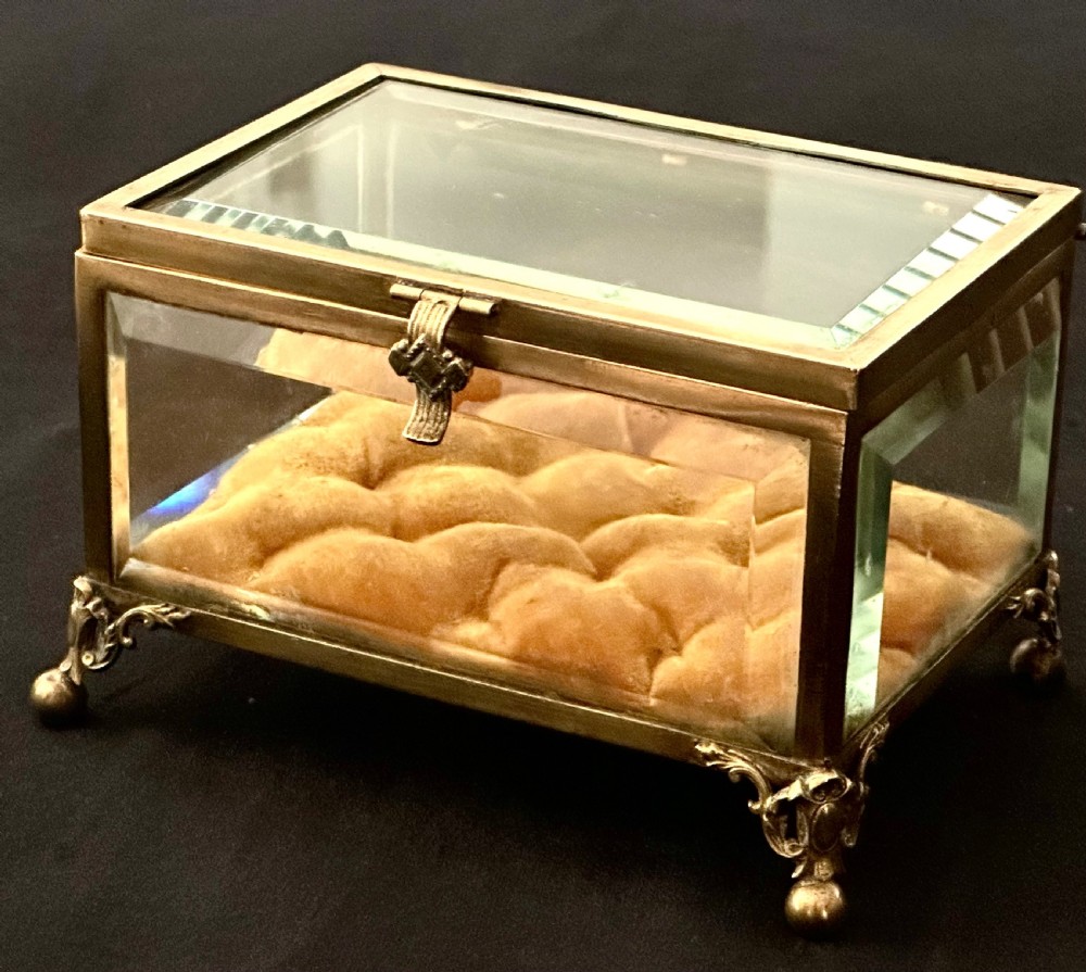 A rectangular French glass trinket box featuring a gilded ormolu frame, bevelled glass panels, decorative cast feet, and a tufted tan velvet cushion inside.