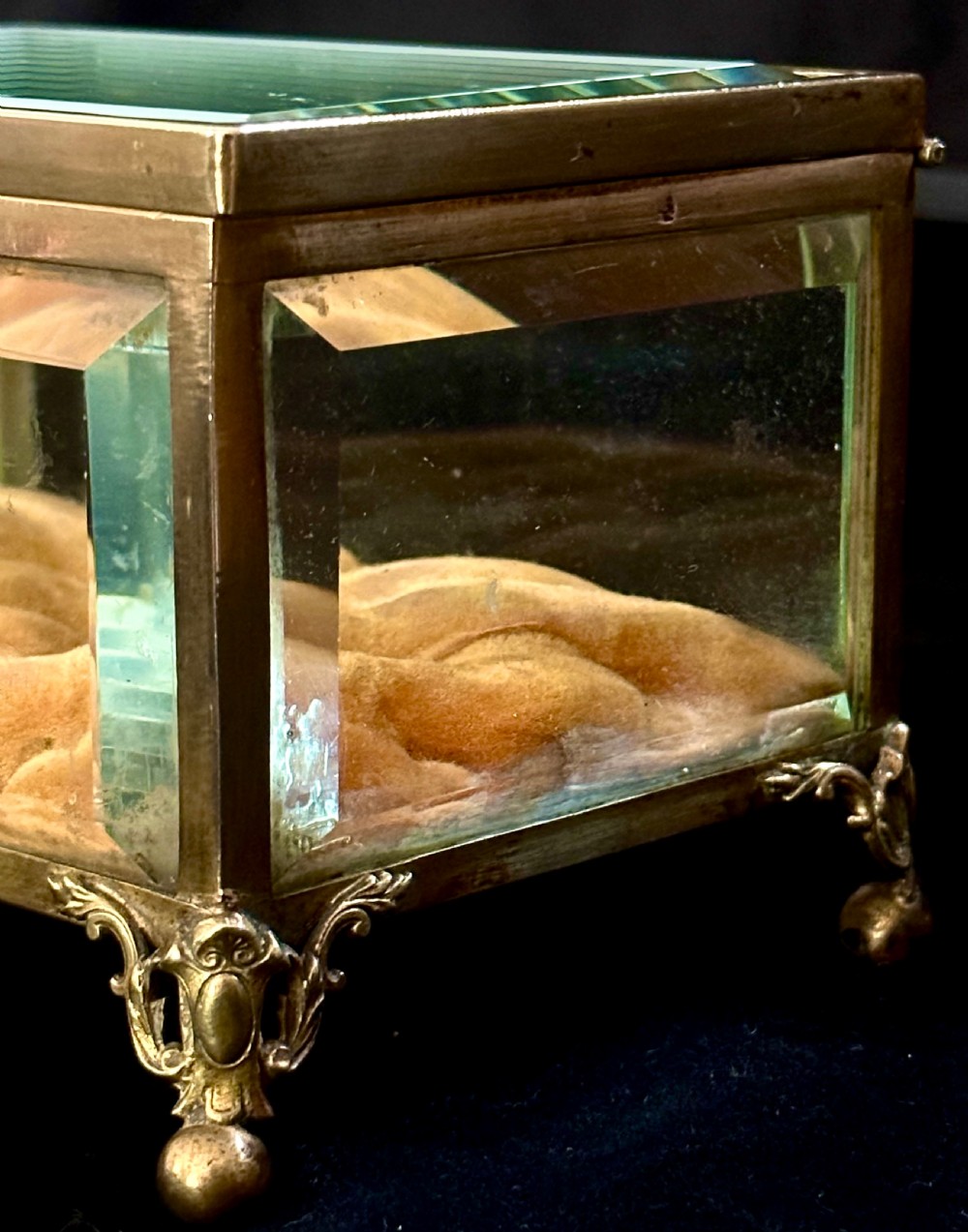 A rectangular French glass trinket box featuring a gilded ormolu frame, bevelled glass panels, decorative cast feet, and a tufted tan velvet cushion inside. View III