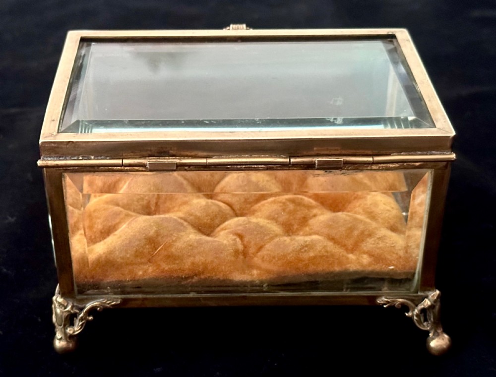 A rectangular French glass trinket box featuring a gilded ormolu frame, bevelled glass panels, decorative cast feet, and a tufted tan velvet cushion inside. View VIII