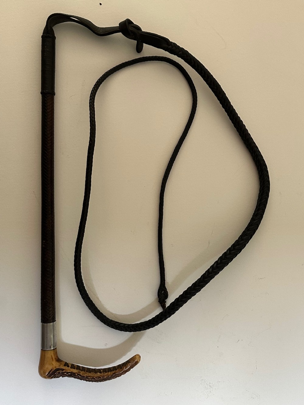 An antique riding crop featuring a carved antler handle, a silver collar, a dark woven shaft, and a braided leather lash.