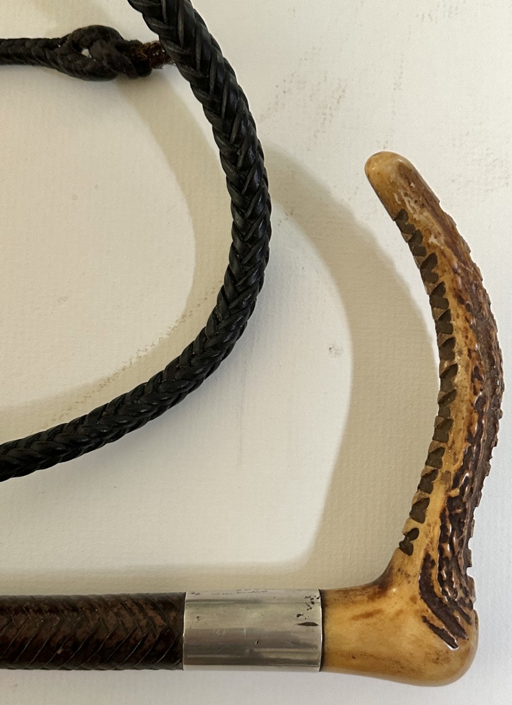 An antique riding crop featuring a carved antler handle, a silver collar, a dark woven shaft, and a braided leather lash. View II