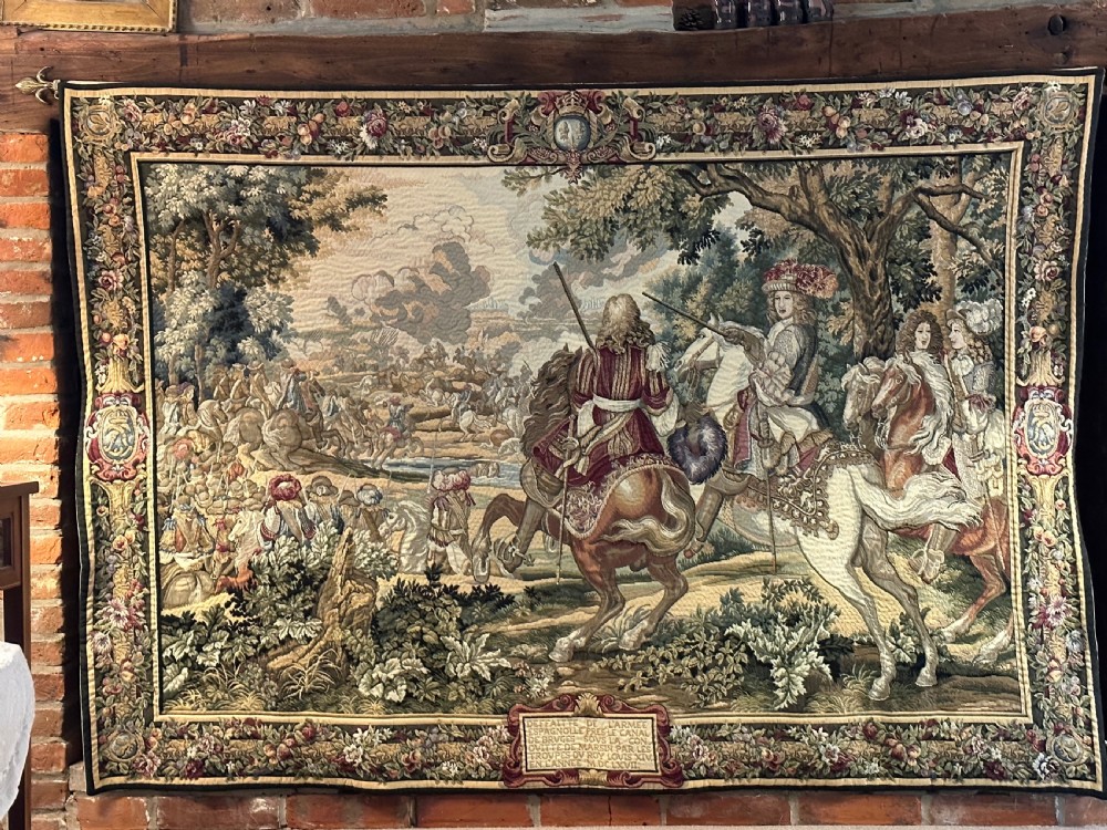 A large needlework wall tapestry featuring a French battle scene with cavalry figures enclosed by a detailed floral border and heraldic crest.