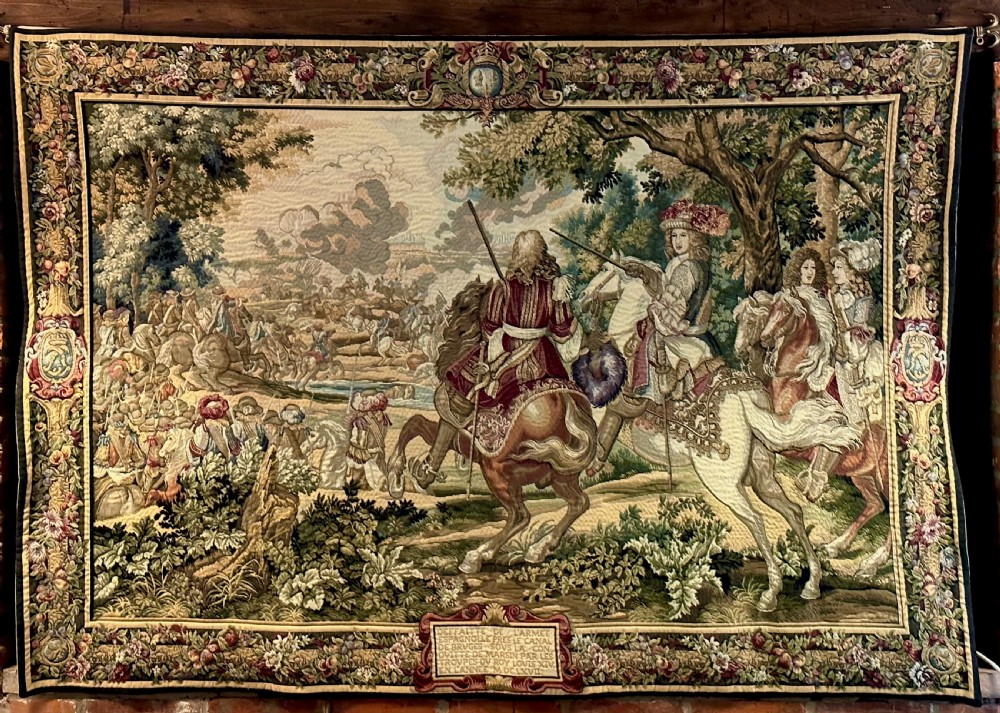 A large needlework wall tapestry featuring a French battle scene with cavalry figures enclosed by a detailed floral border and heraldic crest. View III