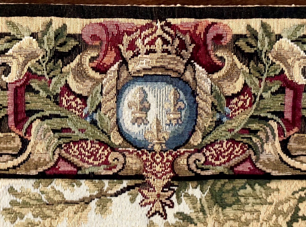 A large needlework wall tapestry featuring a French battle scene with cavalry figures enclosed by a detailed floral border and heraldic crest. View IV