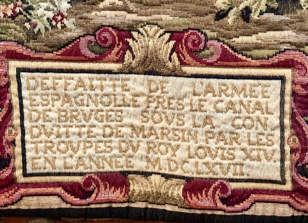 A large needlework wall tapestry featuring a French battle scene with cavalry figures enclosed by a detailed floral border and heraldic crest. View VI