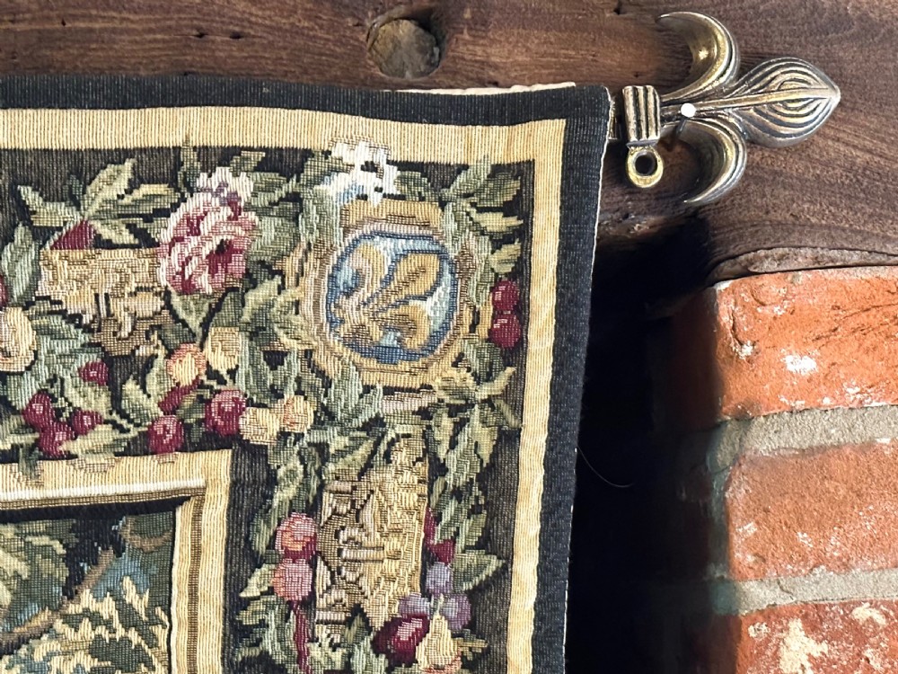 A large needlework wall tapestry featuring a French battle scene with cavalry figures enclosed by a detailed floral border and heraldic crest. View VII