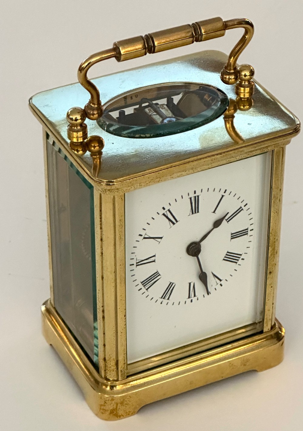 A rectangular brass French carriage clock featuring a white enamel dial with Roman numerals, black hands, bevelled glass sides, and a hinged carrying handle on top. View VI