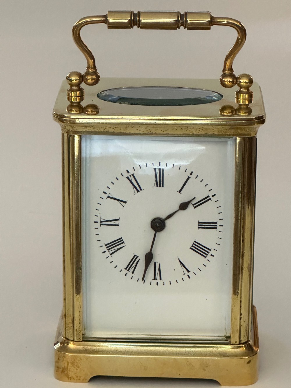 A rectangular brass French carriage clock featuring a white enamel dial with Roman numerals, black hands, bevelled glass sides, and a hinged carrying handle on top. View VIII