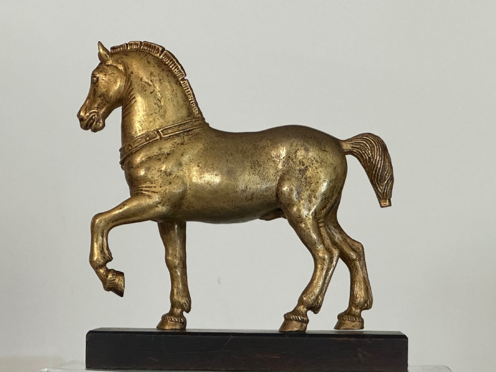A gilded bronze horse sculpture in a striding pose with one raised hoof, mounted on a dark rectangular base. View III