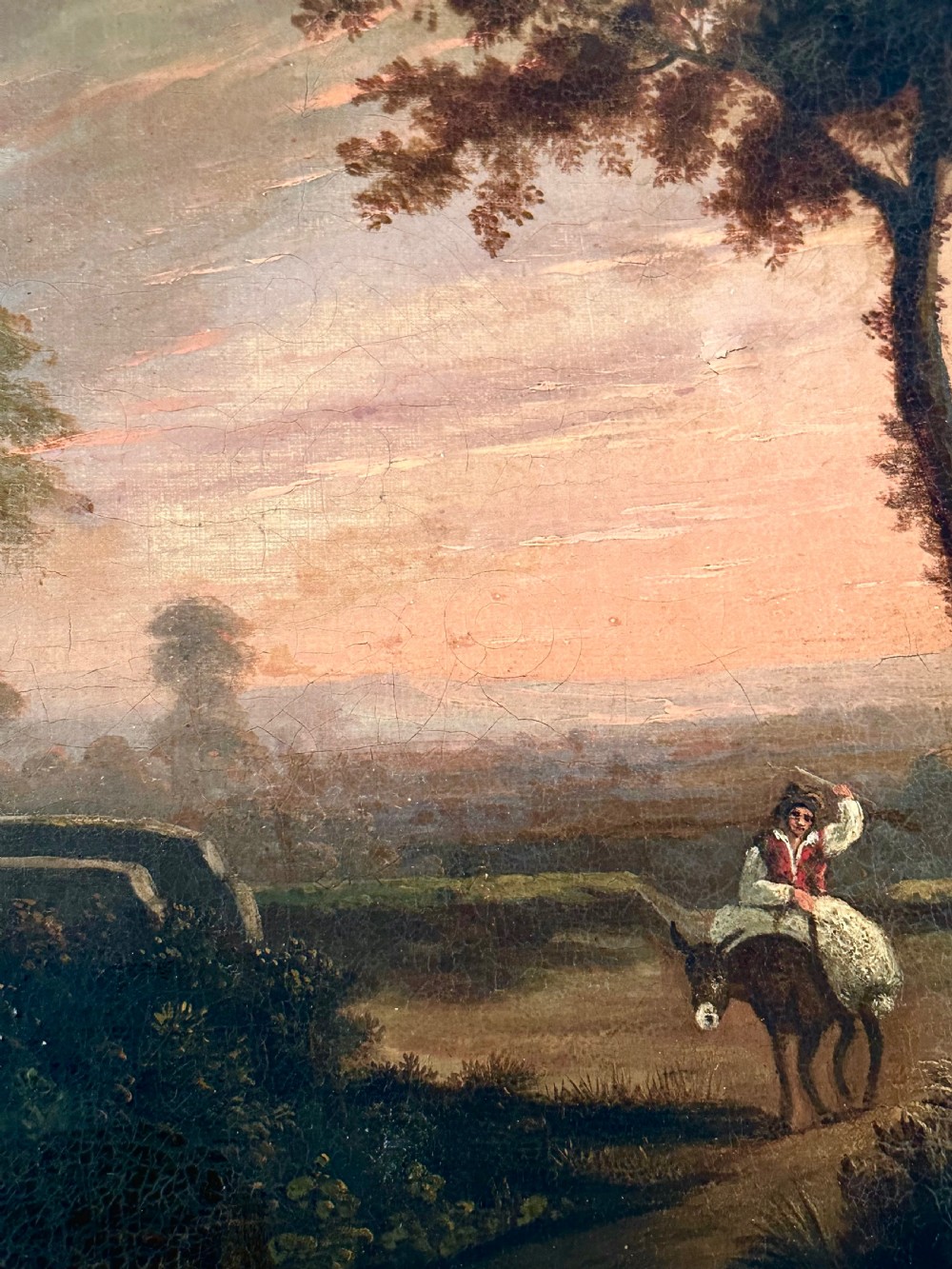 An 18th-century oil painting on canvas depicting a rural landscape with a stone gatehouse, a figure riding a donkey, and a warm sunset sky, set within an ornate carved gold gilt frame. View IV