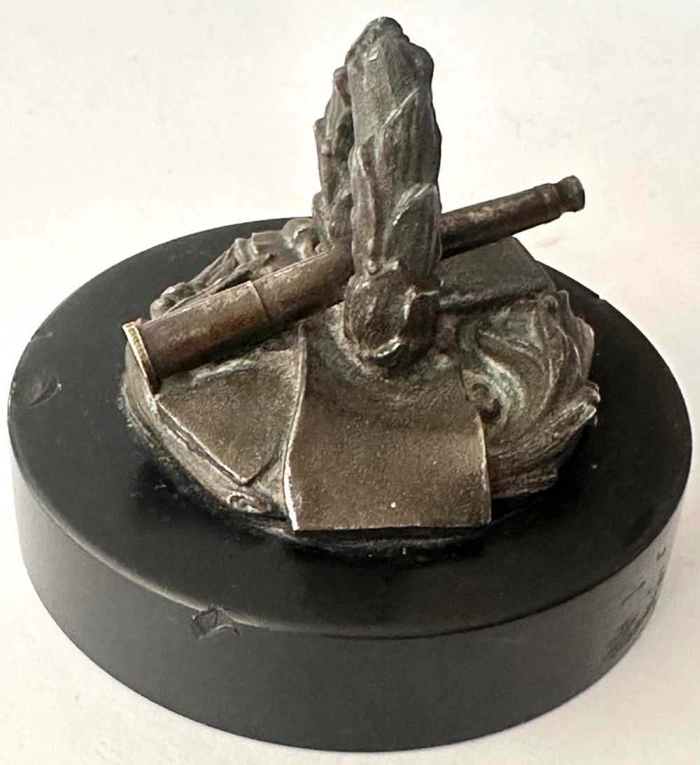 A small bronze military-themed sculpture featuring a laurel wreath, telescope, books, and scroll mounted on a circular black base. View III