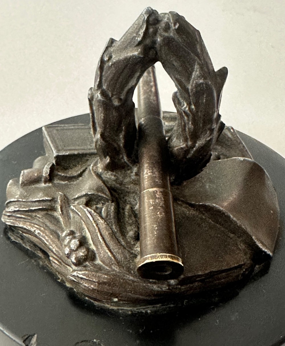 A small bronze military-themed sculpture featuring a laurel wreath, telescope, books, and scroll mounted on a circular black base. View IV