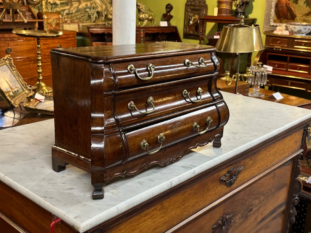 A miniature dark oak Dutch apprentice chest of drawers featuring a bombee front, three drawers with brass drop handles, and carved bracket feet.