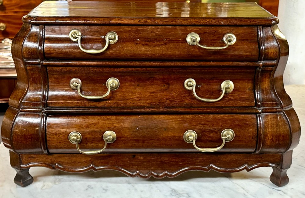 A miniature dark oak Dutch apprentice chest of drawers featuring a bombee front, three drawers with brass drop handles, and carved bracket feet. View II