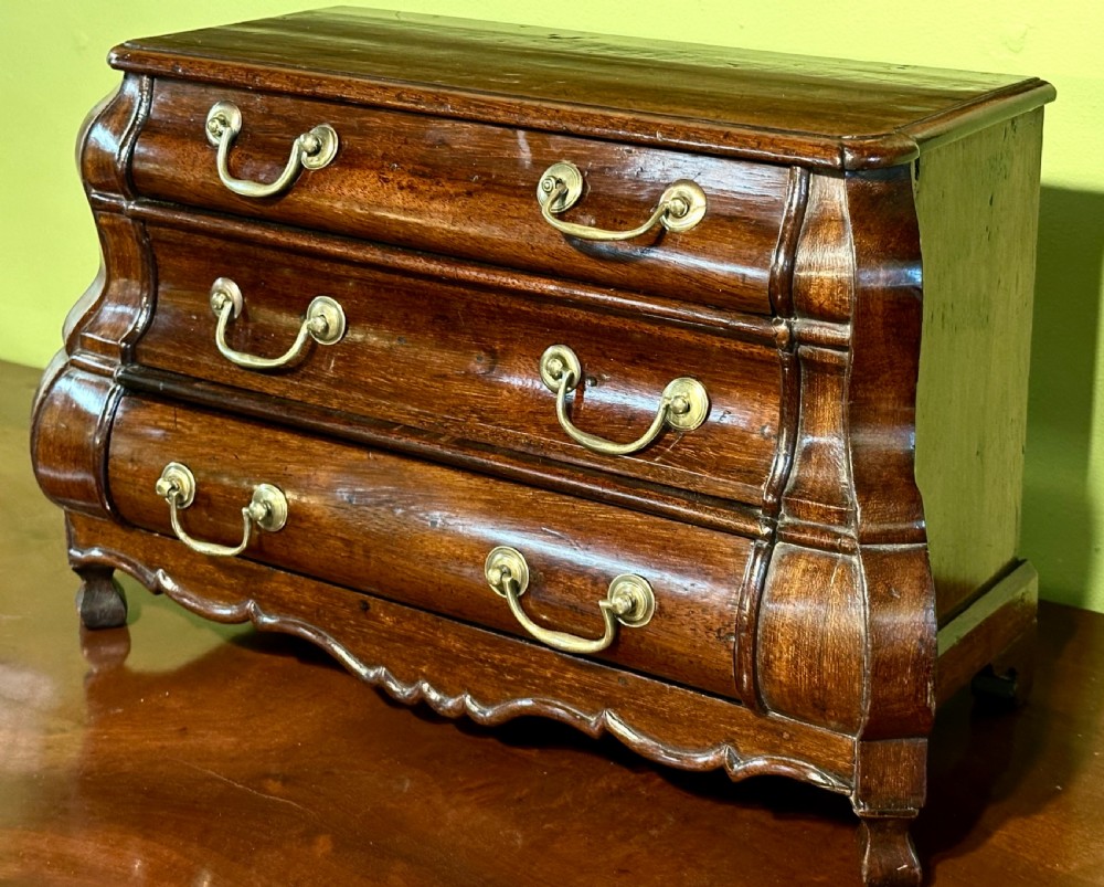 A miniature dark oak Dutch apprentice chest of drawers featuring a bombee front, three drawers with brass drop handles, and carved bracket feet. View IX
