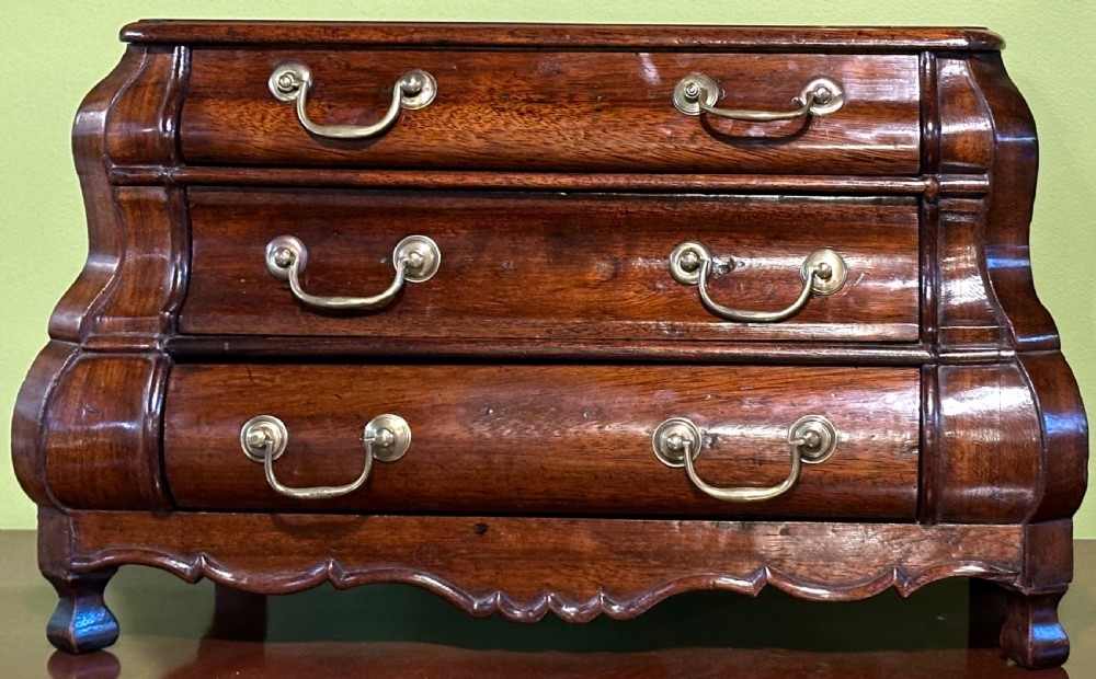 A miniature dark oak Dutch apprentice chest of drawers featuring a bombee front, three drawers with brass drop handles, and carved bracket feet. View X