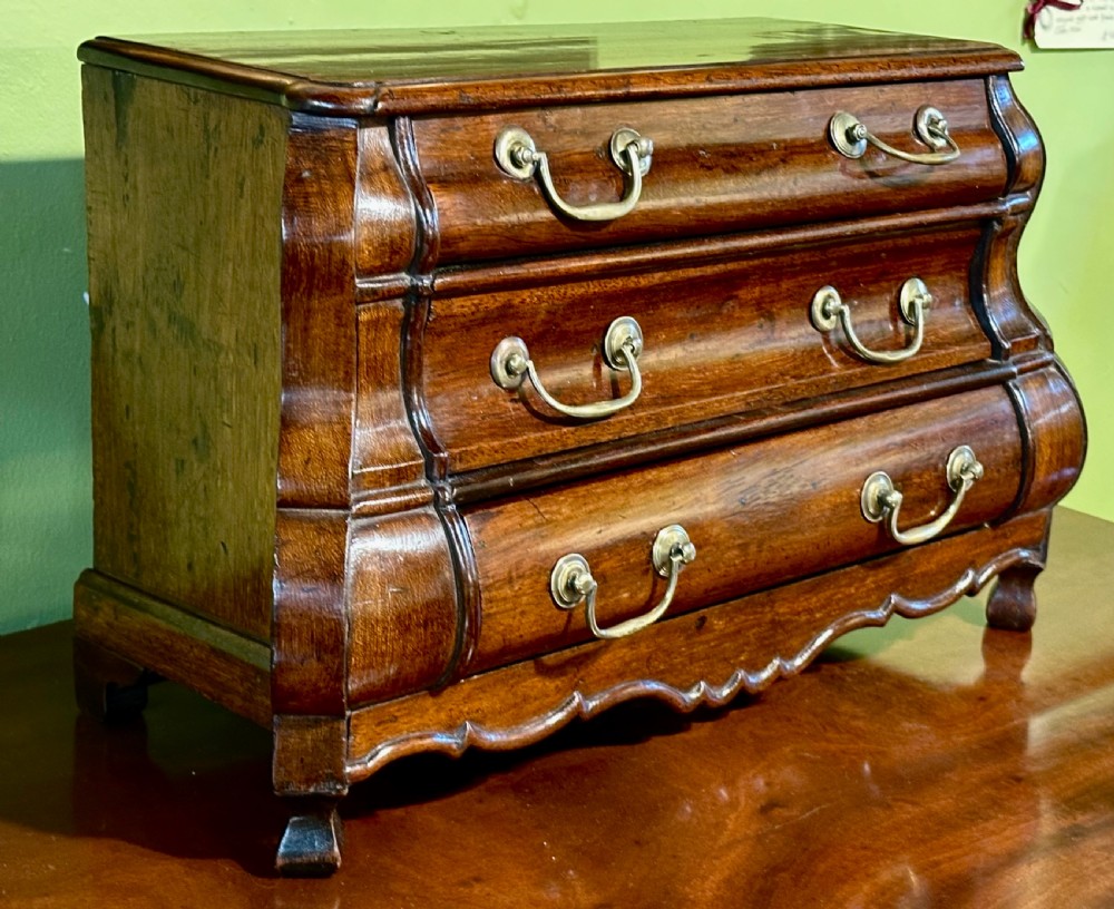 A miniature dark oak Dutch apprentice chest of drawers featuring a bombee front, three drawers with brass drop handles, and carved bracket feet. View XI