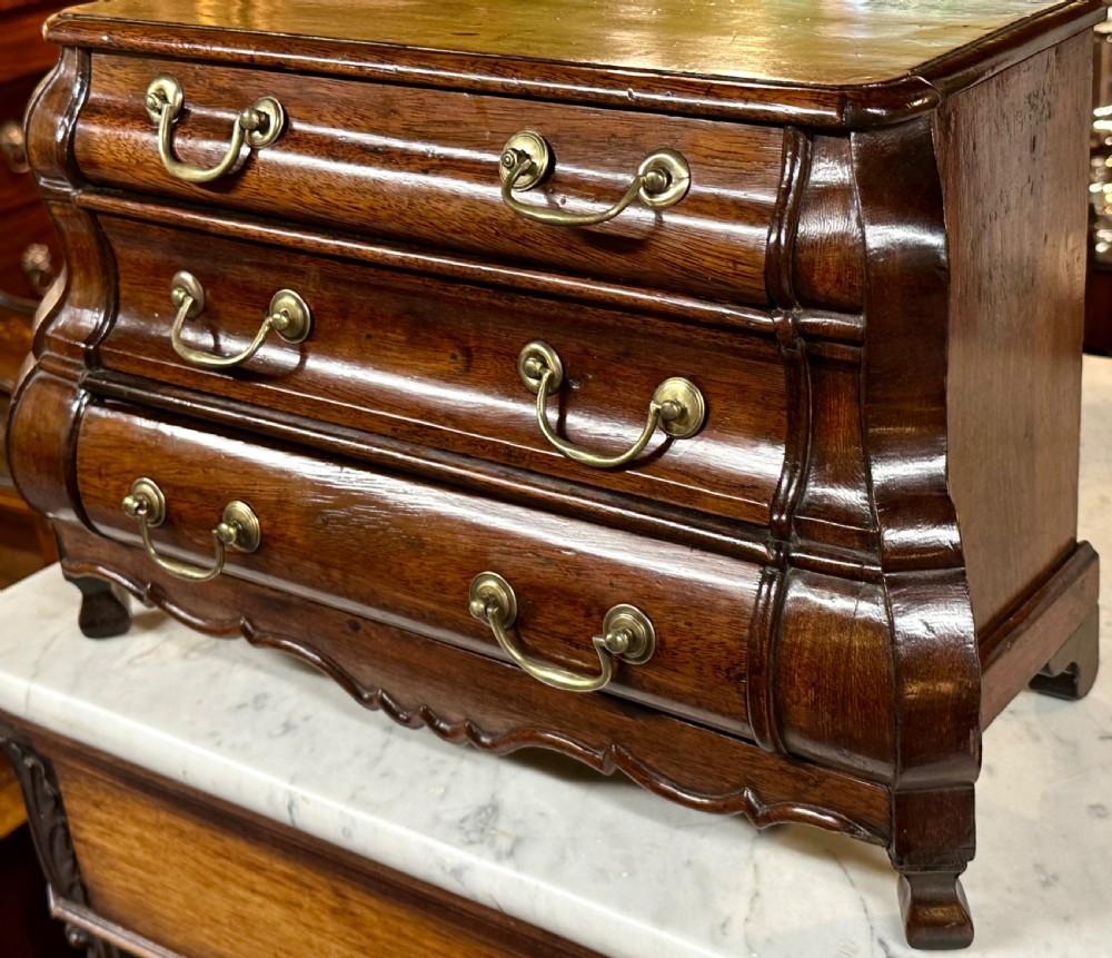 A miniature dark oak Dutch apprentice chest of drawers featuring a bombee front, three drawers with brass drop handles, and carved bracket feet. View XIV
