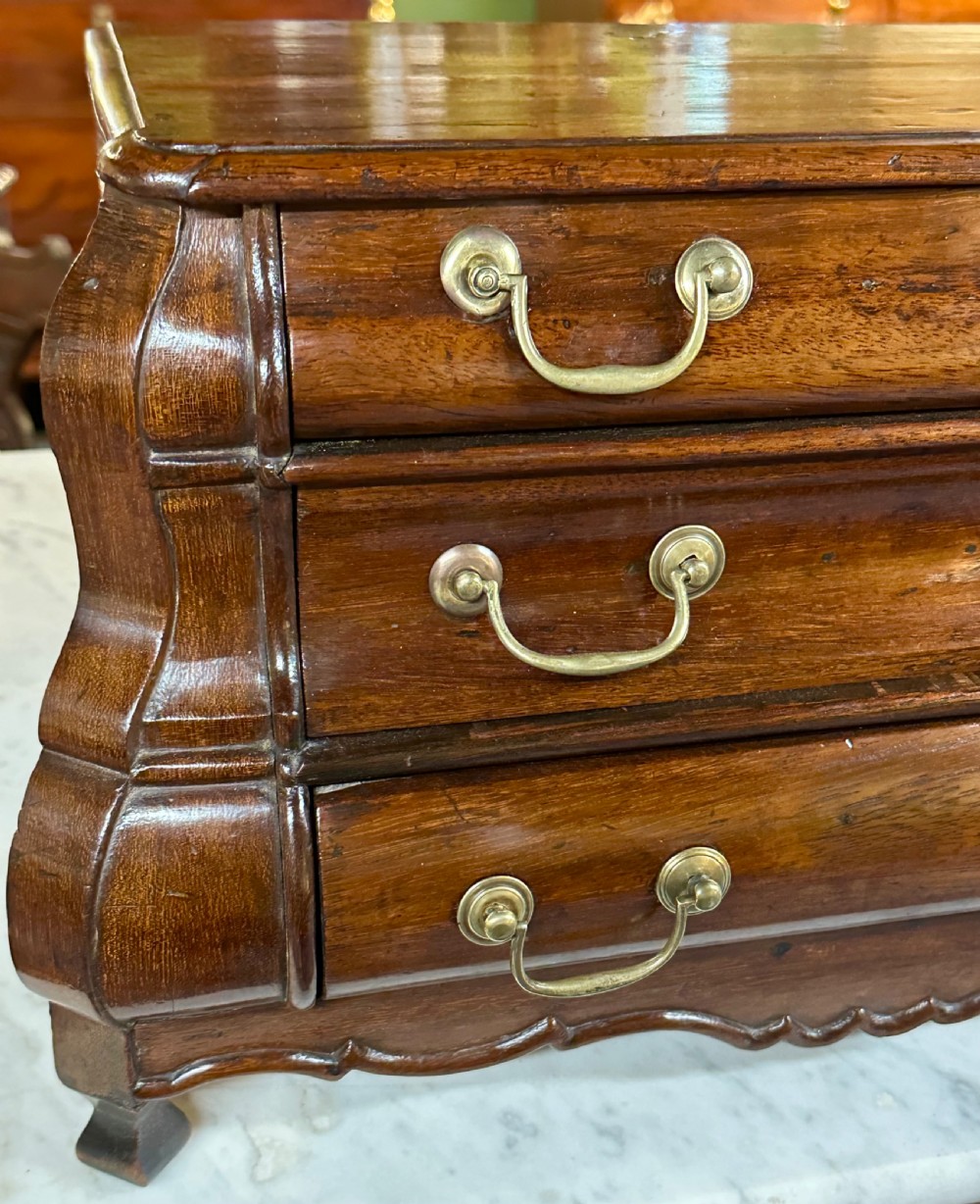 A miniature dark oak Dutch apprentice chest of drawers featuring a bombee front, three drawers with brass drop handles, and carved bracket feet. View XV