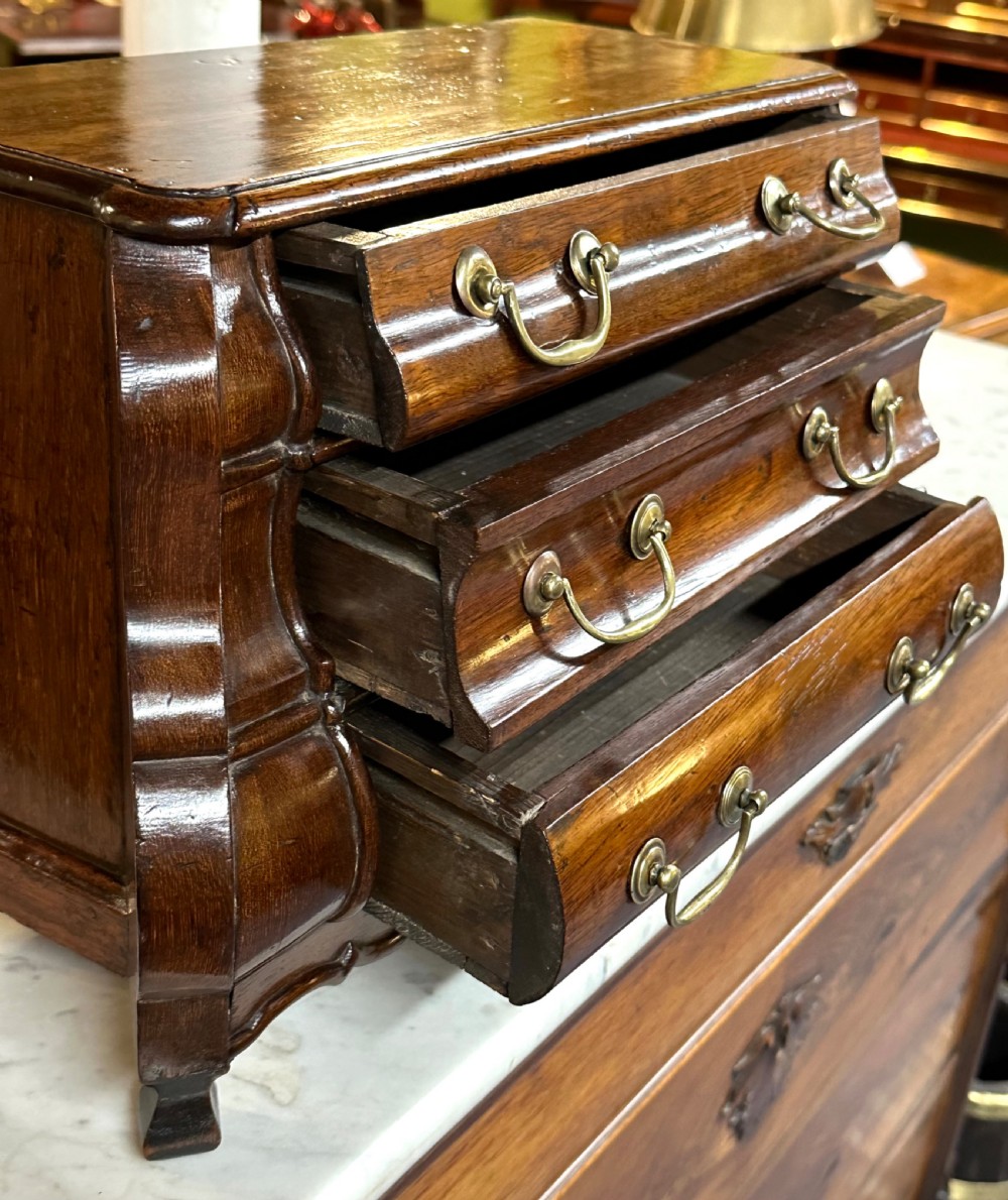 A miniature dark oak Dutch apprentice chest of drawers featuring a bombee front, three drawers with brass drop handles, and carved bracket feet. View XVIII