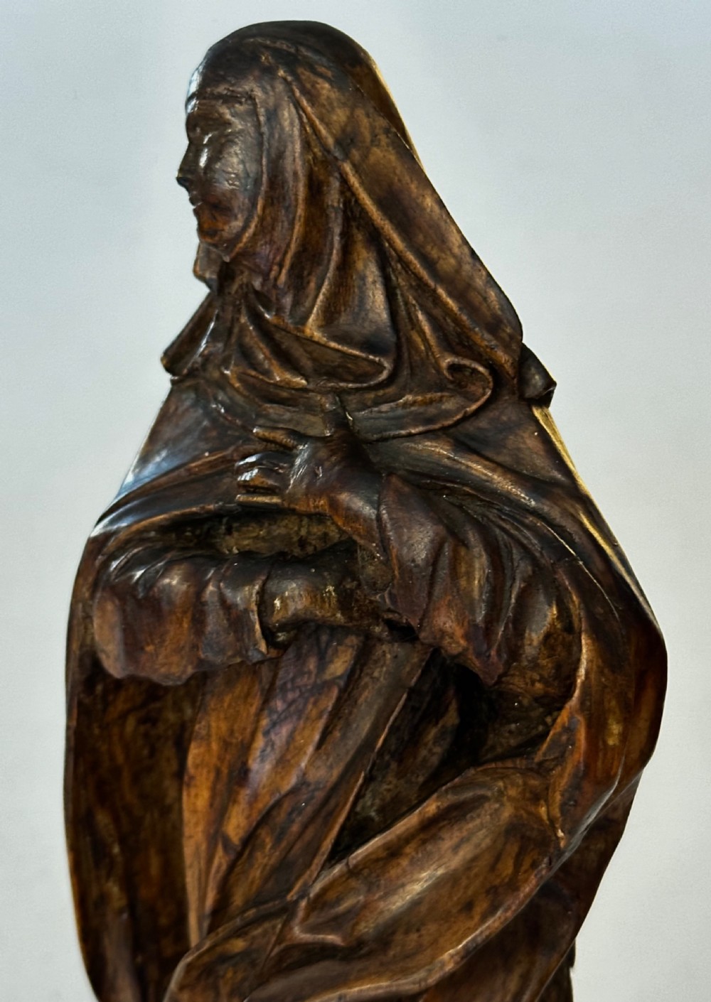 A dark carved wood statue depicting a veiled female religious saint with draped clothing on a faceted wooden base. View III