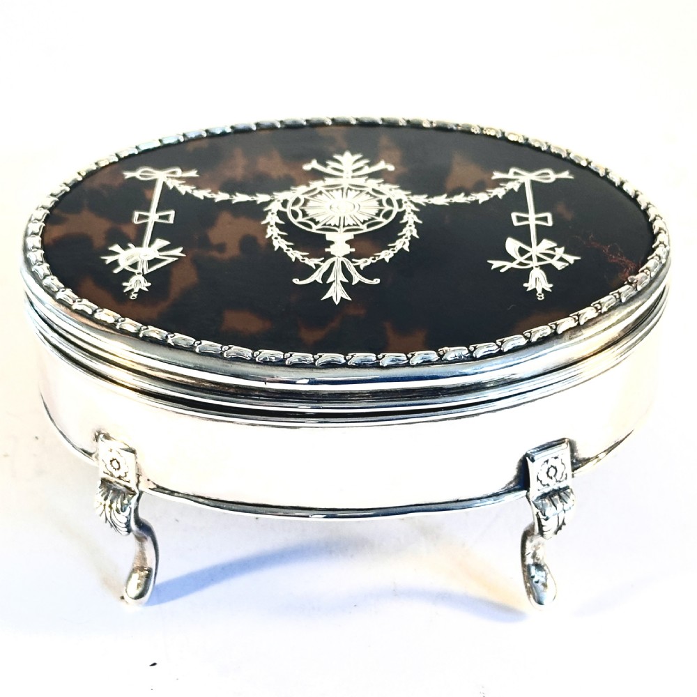 An oval sterling silver jewellery box raised on four small legs, featuring a dark brown tortoiseshell lid inlaid with silver swags and ribbon motifs.