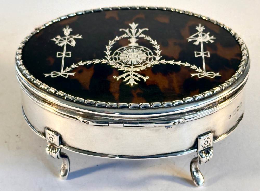An oval sterling silver jewellery box raised on four small legs, featuring a dark brown tortoiseshell lid inlaid with silver swags and ribbon motifs. View II