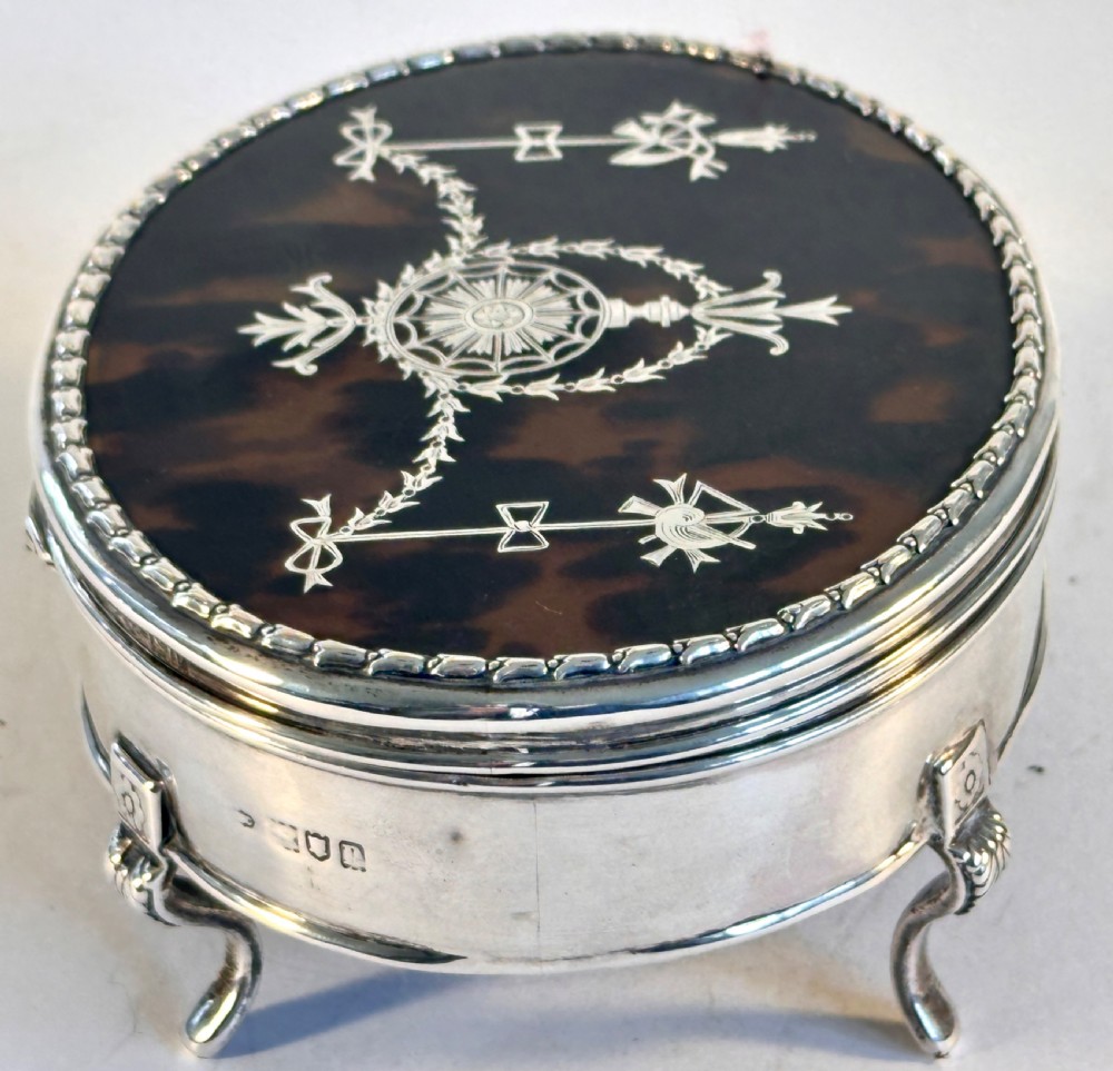An oval sterling silver jewellery box raised on four small legs, featuring a dark brown tortoiseshell lid inlaid with silver swags and ribbon motifs. View III