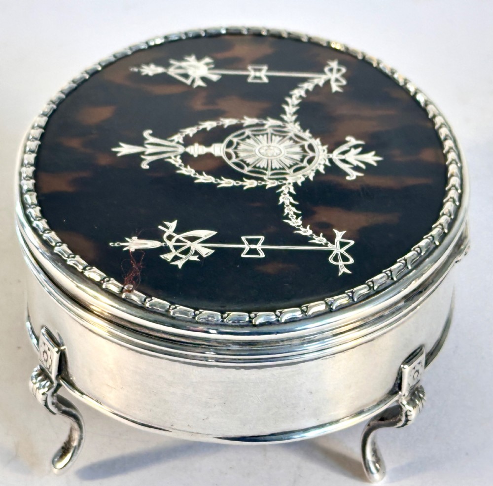 An oval sterling silver jewellery box raised on four small legs, featuring a dark brown tortoiseshell lid inlaid with silver swags and ribbon motifs. View IV