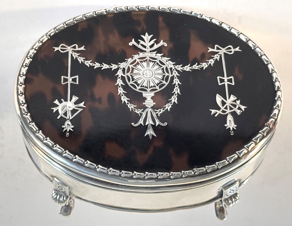An oval sterling silver jewellery box raised on four small legs, featuring a dark brown tortoiseshell lid inlaid with silver swags and ribbon motifs. View V