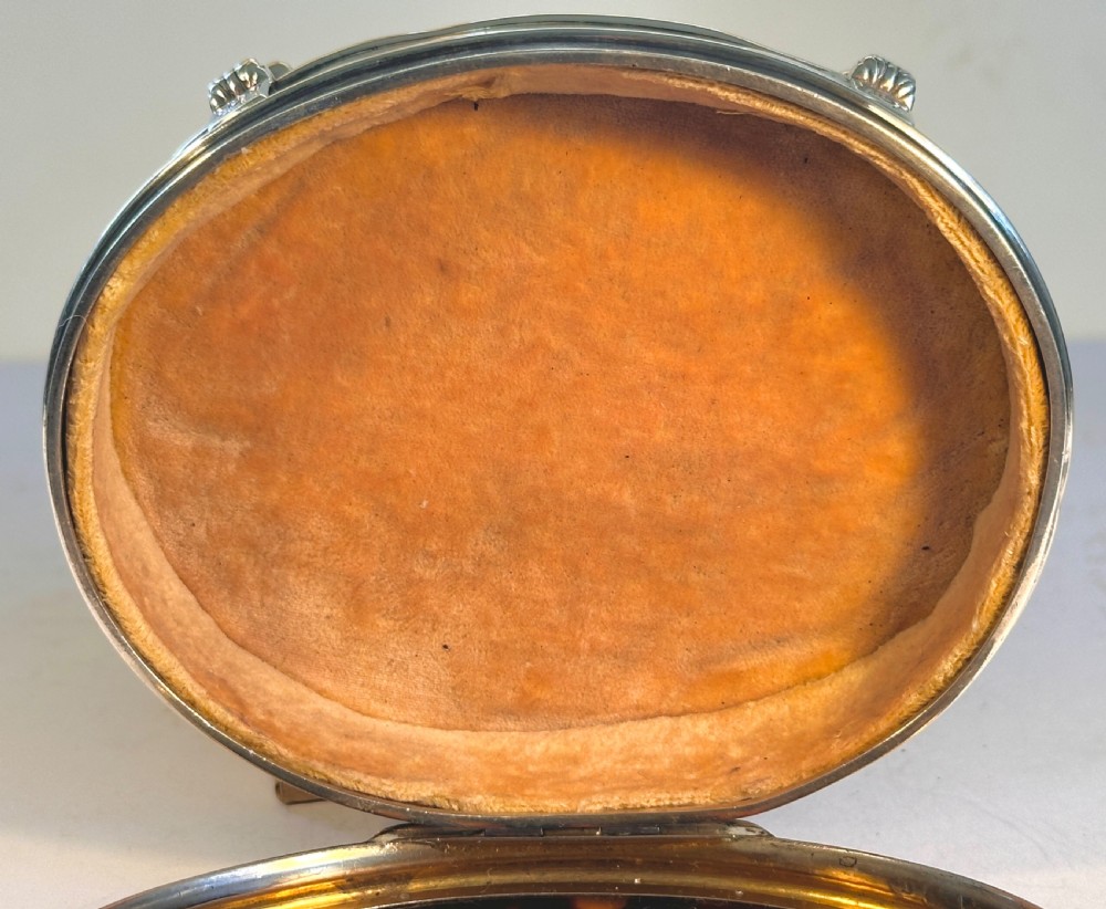 An oval sterling silver jewellery box raised on four small legs, featuring a dark brown tortoiseshell lid inlaid with silver swags and ribbon motifs. View XI
