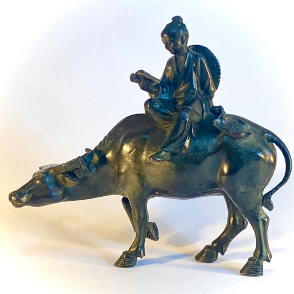 A dark patinated bronze sculpture featuring a scholar seated atop a water buffalo while reading an open book.