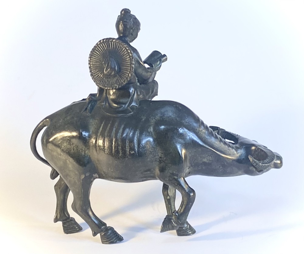 A dark patinated bronze sculpture featuring a scholar seated atop a water buffalo while reading an open book. View II