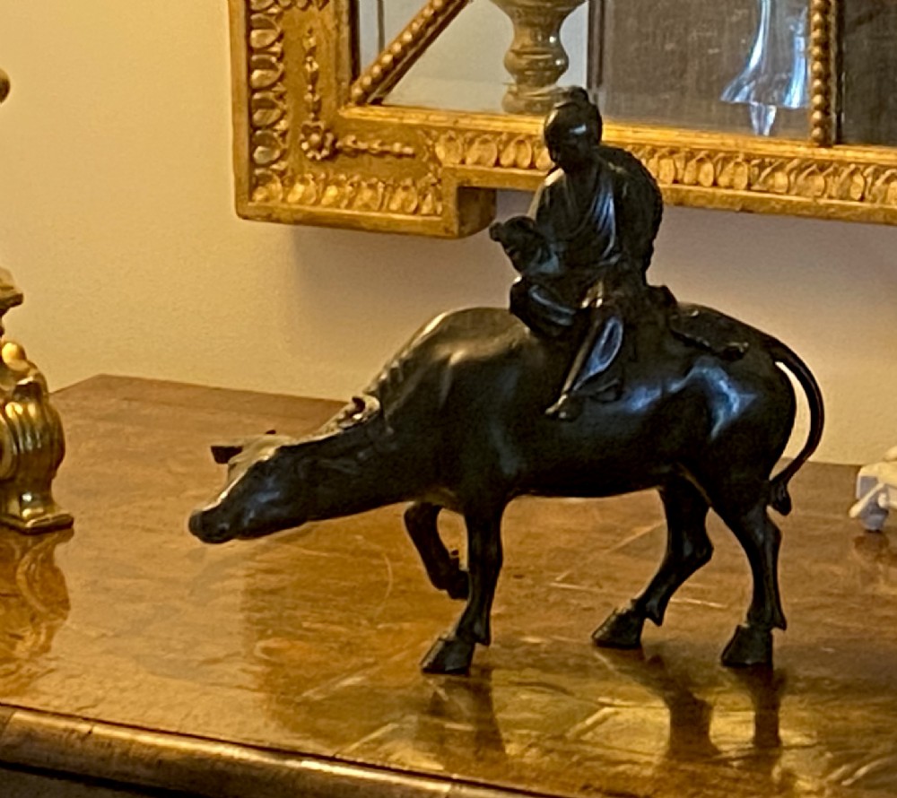 A dark patinated bronze sculpture featuring a scholar seated atop a water buffalo while reading an open book. View XI