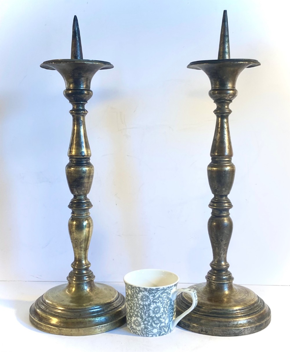 A pair of bronze church pricket candlesticks featuring turned baluster stems, circular stepped bases, drip trays, and central spikes. View VI