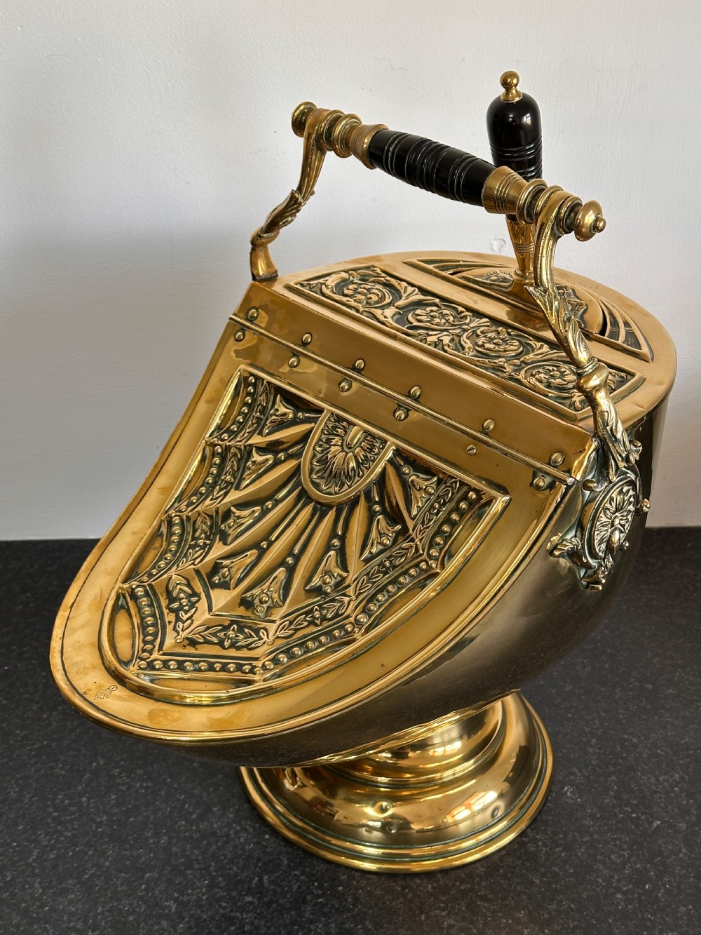 Victorian brass coal scuttle featuring elaborate repousse relief patterns, a pedestal base, and an ebonised wooden handle.