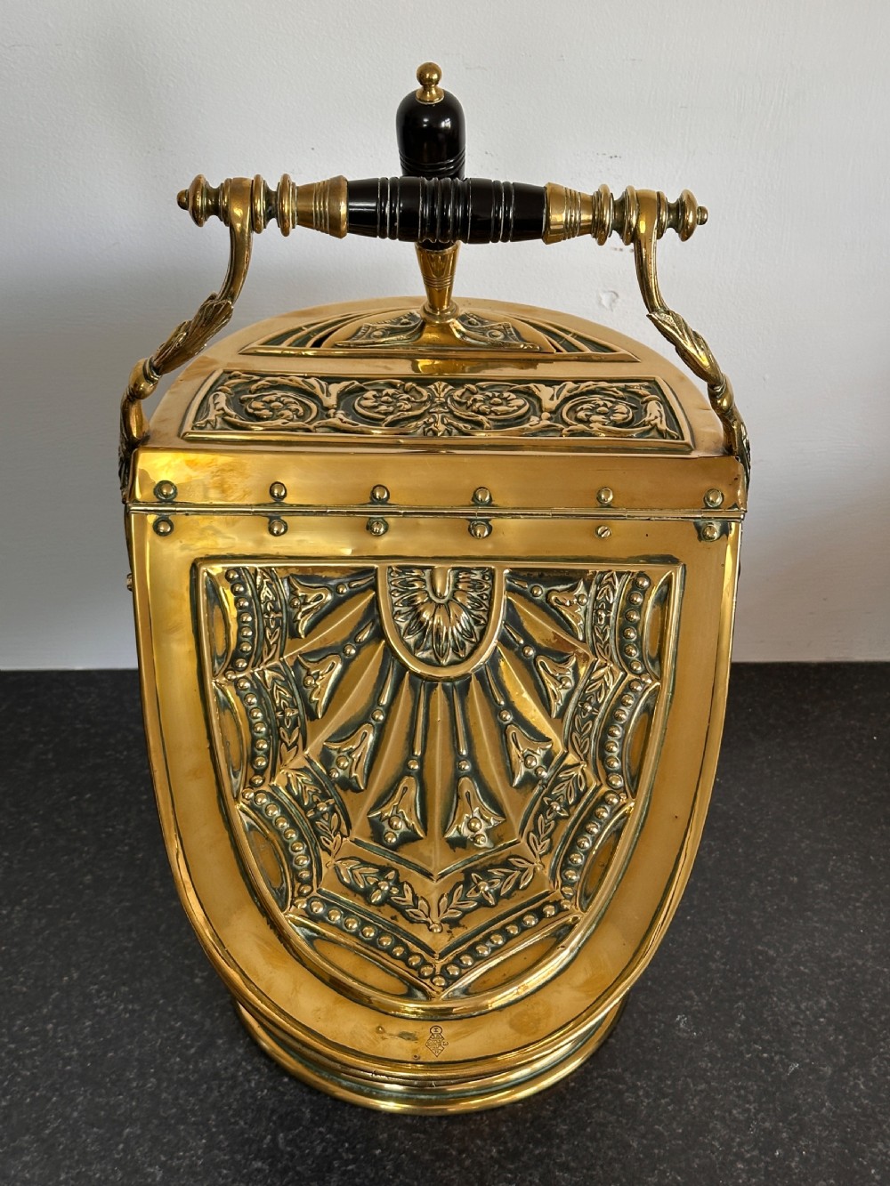 Victorian brass coal scuttle featuring elaborate repousse relief patterns, a pedestal base, and an ebonised wooden handle. View V