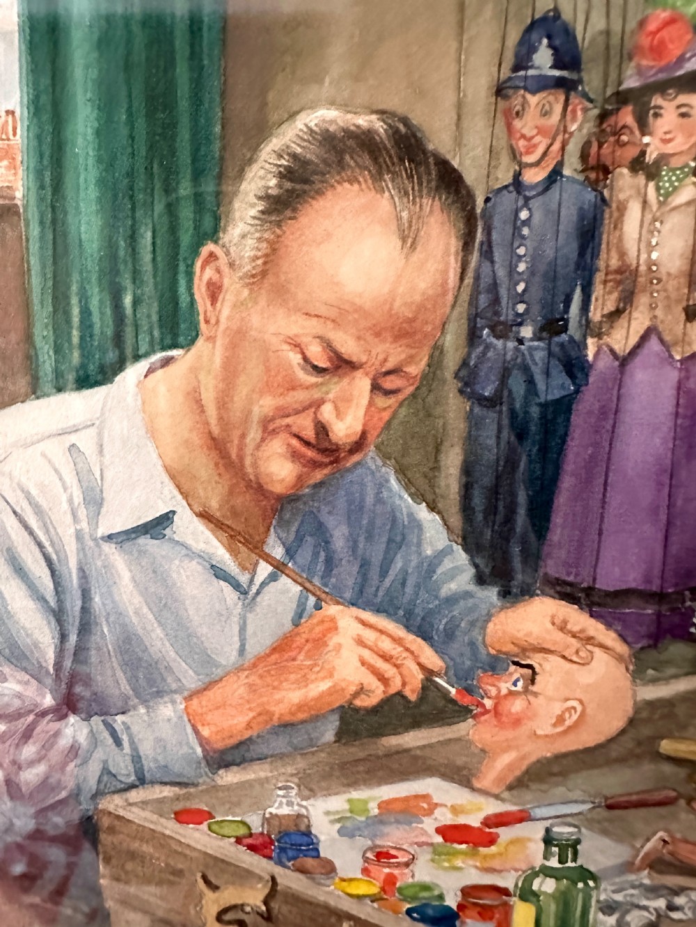 A framed watercolour painting depicting a craftsman at a workbench painting a marionette head, surrounded by finished puppets hanging in the background. View III