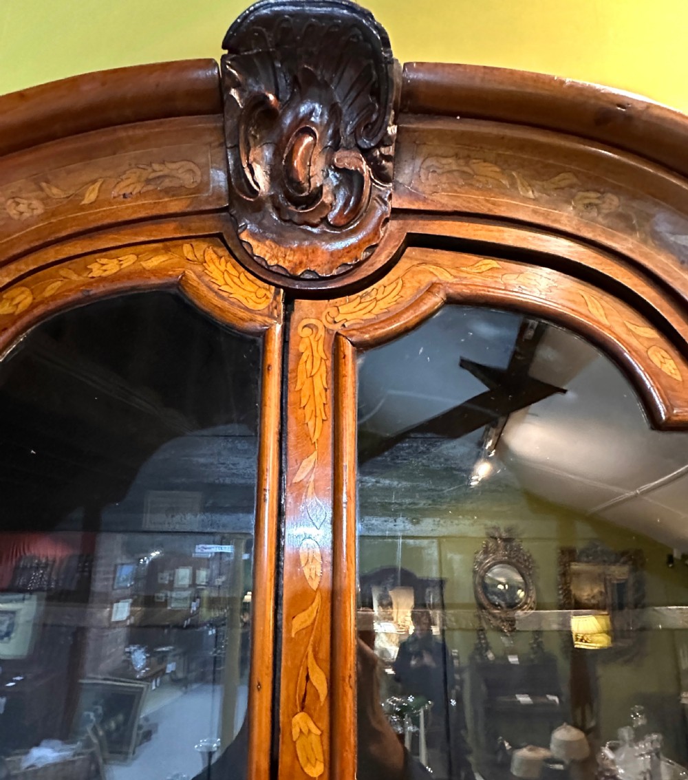 An 18th-century Dutch walnut display cabinet featuring a shaped bonnet top, glazed doors, intricate floral marquetry, and a lower bombe chest with drawers. View VI